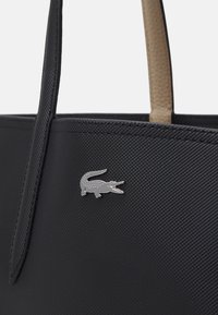 Czarna torba typu tote z teksturowanego materiału z beżowymi uchwytami i srebrnym akcentem w postaci logo krokodyla z przodu. Wytrzymały materiał o eleganckim designie.