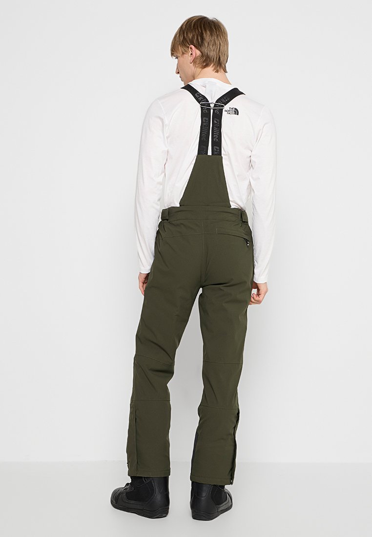 Mann steht mit dem Rücken zur Kamera, trägt olivgrüne Latzhose, ein weißes Langarmshirt, schwarze Stiefel und schwarze Hosenträger vor einem schlichten weißen Hintergrund.