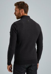 Maglione di lana lavorato a maglia di colore grigio scuro, con collo alto, motivo testurizzato e polsini e orlo a coste. Include una toppa sul maniche sinistro.