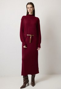 Rochie de pulover lungă, burgundy, cu mânecă lungă, guler înalt și talie ajustată, decorată cu un curea subțire maro, textură simplă și lungime până la genunchi.