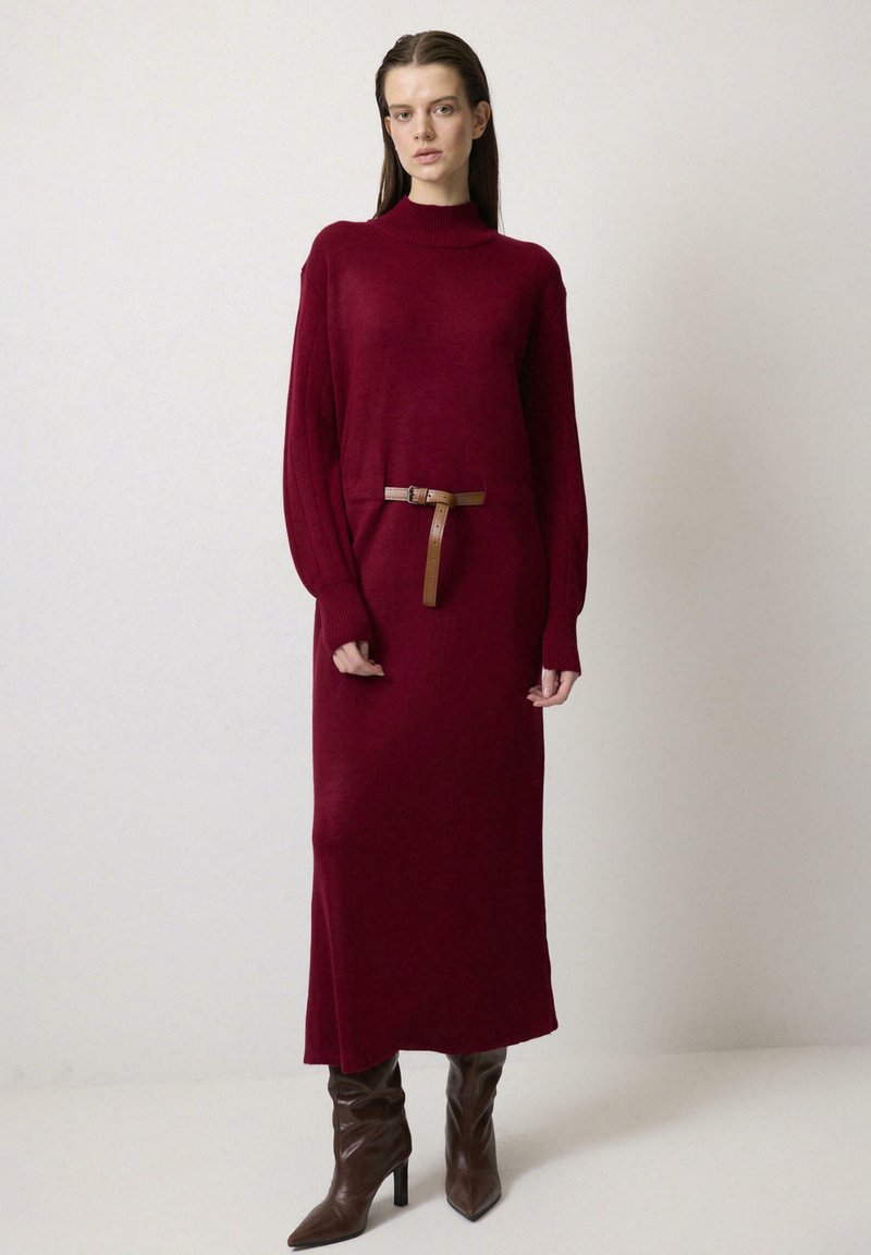 Rochie de pulover lungă, burgundy, cu mânecă lungă, guler înalt și talie ajustată, decorată cu un curea subțire maro, textură simplă și lungime până la genunchi.