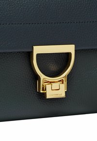 Borsa a mano in pelle blu scuro con texture, dotata di una chiusura rettangolare dorata e un manico circolare, nome del marchio inciso sull'hardware.