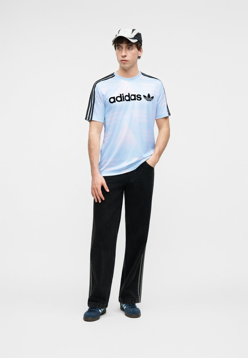 Junger Mann, der ein hellblaues Adidas-T-Shirt mit schwarzen Streifen, schwarze Hosen, blaue Sneakers und eine weiß-schwarze Kappe trägt, steht und schaut nach rechts.