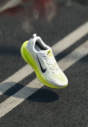 Weißer und gelber Nike-Laufschuh mit "ZoomX"-Sohle in der Luft über einer Straße mit weißen Fahrbahnmarkierungen.