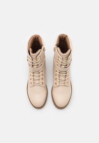 Pár béžových kožených kotníkových bot s tkaničkami, bočními zipy, polstrovanými límci a hnědými gumovými podrážkami na bílém pozadí.