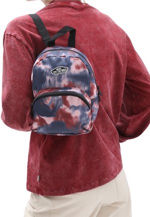 Persona con una pequeña mochila tie-dye en colores azul marino, rojo y rosa, sobre una camisa roja de manga larga y pantalones beige.