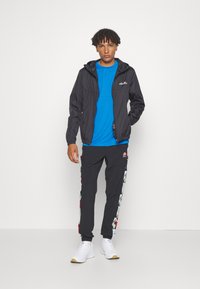 Schwarze Windjacke, blaues T-Shirt, schwarze Hose mit mehrfarbigen seitlichen Akzenten, weiße Sneakers, stehend vor einem einfarbigen Hintergrund.