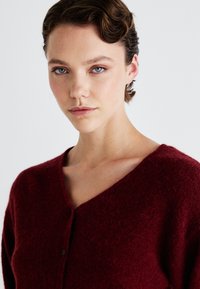 Burgundy knitted cardigan with a V-neck, featuring a button closure and a soft, textured finish. Simple design without patterns or accents.