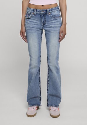 Persona che indossa jeans bootcut blu sbiaditi e sneakers rosa chiaro, in piedi su uno sfondo semplice.