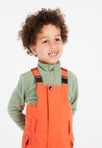 Barnet har en orange vattentät overall med justerbara svarta remmar, kombinerad med en grön fleece-tröja. Hen ler och visar upp ett gapande leende.