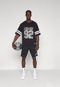 T-shirt oversized preta com impressão em prata de "Champion" e "92", combinada com calções pretos, ténis brancos e segurando uma bola de basquetebol.