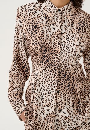 Dopasowana koszula zapinana na guziki z beżowym, brązowym i czarnym nadrukiem w panterę, noszona z rękami w kieszeniach.