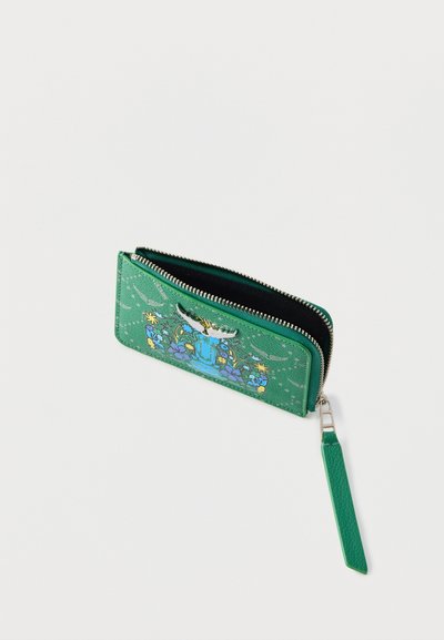 Πράσινο πορτοφόλι με φερμουάρ και λουρί χειρός, σχέδιο με λουλούδια και πουλιά, και ασημένιο έμβλημα πουλιού σε υφασμένη επιφάνεια.