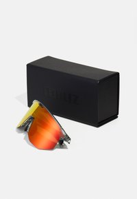 Sportsonnenbrille mit großen orangen Verlaufsgläsern, einem klaren Rahmen und metallischen Akzenten, platziert neben einer schwarzen Box mit Logo.