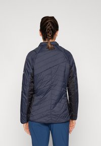 Marinblå puffjacka med en chevronquiltad design, långa ärmar och hög krage. Har kontrasterande svarta sidor och en jämn yta.