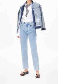 Chaqueta de mezclilla con un lavado azul claro, detalles en azul oscuro y estampado; camisa blanca de botones debajo; jeans rectos azul claro; sandalias de plataforma negras.