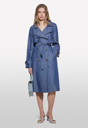 Blauer Denim-Trenchcoat mit doppelt geknöpften Verschluss, tailliertem Gürtel, breiten Revers und Seitentaschen; Modell hält eine hellblaue Tasche.