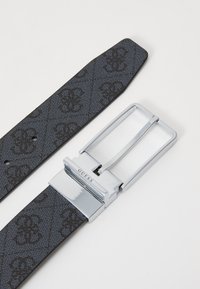 Svart strukturert belte med preget mønster, med en sølvmetallspenne gravert med "GUESS." Beltet smalner av i den ene enden.