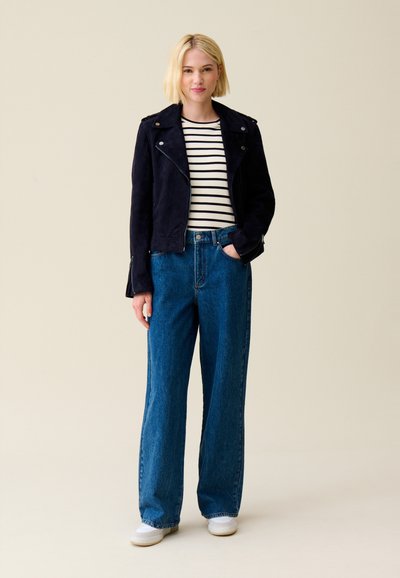 Veste de motard en cuir suédé bleu marine sur une chemise rayée blanche et noire, associée à un jean en denim bleu à jambes larges et des baskets blanches.