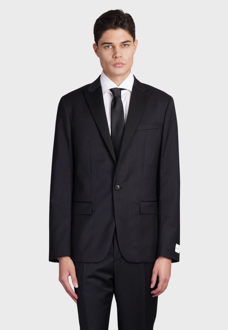 Completo nero con rever satinato, chiusura a un bottone, due tasche frontali e design elegante. Include una camicia bianca e una cravatta nera.