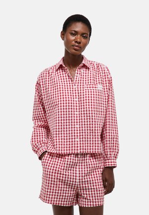 Femme portant une chemise à manches longues à carreaux rouges et blancs boutonnée et un short assorti, debout les mains dans les poches devant un fond uni.