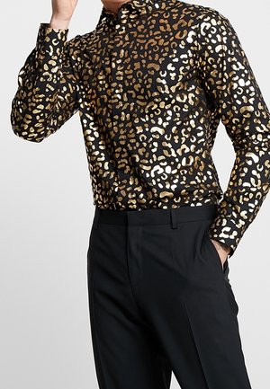 Mand iført en skinnende sort skjorte med brunt og gyldent leopardprint, kombineret med sorte bukser, hånd i lommen.