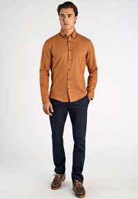 Brązowa koszula z guzikami, wykonana z teksturowanej tkaniny, ciemnogranatowe spodnie oraz brązowe buty sznurowane z kontrastowym haftem. Prosty design.