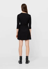 Robe noire à manches trois-quarts, ceinture à la taille, jupe évasée, tissu texturé. Associée à des bottines noires, dos tourné à la caméra.