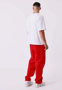Hombre con cabello corto, vistiendo una camiseta blanca, pantalones sudadera rojos, zapatillas blancas y un collar de cadena de plata, de pie de espaldas sobre un fondo liso.
