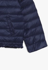 Mörkblå puffjacka med horisontell sömnad, sidficka och rynkad nederkant. Tillverkad av glänsande, lätt material.