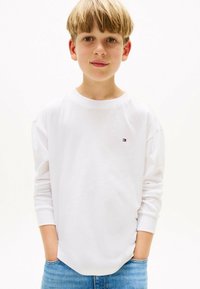 Weißes Langarm-T-Shirt mit rundem Halsausschnitt, mit einem kleinen Logo-Patch auf der linken Brust. Kombiniert mit hellblauen Jeans.