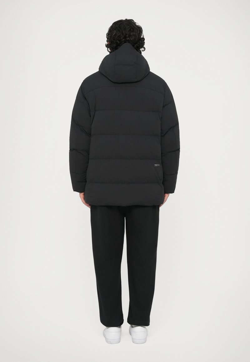 Persona de espaldas vistiendo una chaqueta con capucha negra acolchada, pantalones negros y zapatillas blancas, en un fondo blanco liso.