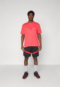 Rød bomulds t-shirt med Kansas City Chiefs logo, parret med sorte shorts med røde detaljer. Båret med grå sokker og sorte sportsko.