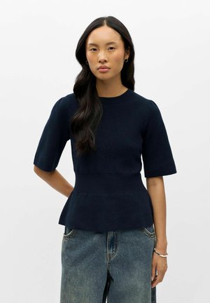 Mujer con cabello largo y oscuro, vestida con una camiseta de punto azul marino de manga corta y vaqueros azules holgados, está de pie frente a un fondo blanco liso.