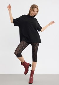 Svart oversized t-skjorte, svarte blondeleggings og røde ankelstøvler med blokkhels. Modellen poserer med armene hevet og ett ben bøyd.