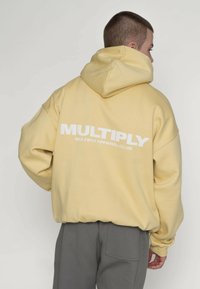 Gele hoodie met een grote witte "MULTIPLY"-logo op de achterkant. Losse pasvorm, zacht materiaal, met een capuchon met trekkoord en doorgezakte schouders.