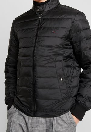 Schwarze gesteppte Jacke mit Reißverschluss und hohem Kragen, seitliche Druckknopftaschen, kleines rot-weiß-blaues Logo auf der Brust, getragen mit grauen karierten Hosen.