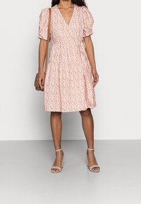 Kvinna iklädd en rosa blommig omslagsklänning med puffärmar, nakna ankelsandaler med remmar, och bärande på en liten brun axelväska.