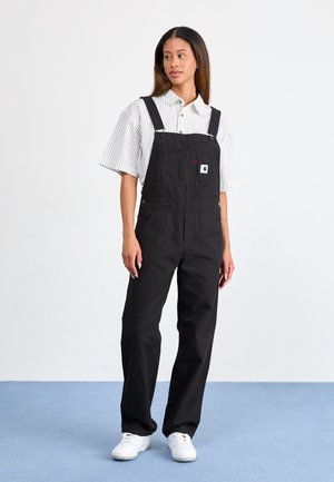 Pantalones de algodón negros con tirantes ajustables, un bolsillo frontal y botones laterales, llevados sobre una camiseta de manga corta a rayas en blanco y gris.
