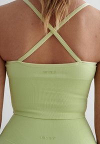 Lys grønn ribbestrikket crop-top med kryssede stropper, med en subtil merke-logo på baksiden. Glatt tekstur og tettsittende design.