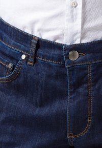 Jeans de mezclilla azul oscuro con cierre de botón, accesorios plateados y costuras en contraste de color naranja. Bolsillos frontales visibles con un ligero detalle de textura.