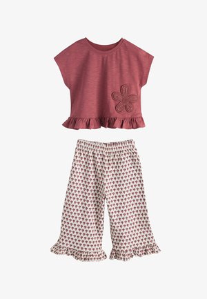 Dijete stoji, nosi bordo kratku majicu s naboranim rubom i ukrasom u obliku cvijeta, široke hlače s uzorkom i sandale u boji ružičastog zlata.