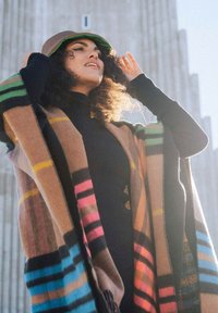 Cape en laine marron avec des rayures multicolores en rose, vert, bleu et jaune. Associé à un chapeau seau marron, porté en plein air contre un fond lumineux.