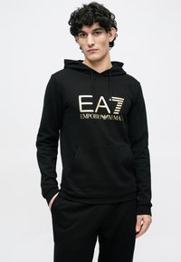 Mladý muž s kučeravými vlasmi stojí s rukami vreckách, oblečený v čiernej mikine s kapucňou so zlatým logom EA7 Emporio Armani na hrudi.