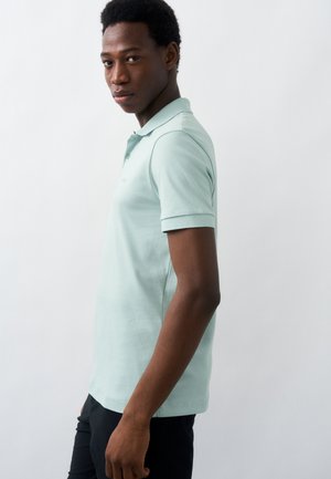 Νέος άνδρας με κοντά σγουρά μαλλιά, φοράει ανοιχτοπράσινο κοντομάνικο πόλο μπλουζάκι και σκούρο παντελόνι, στέκεται πλευρικά μπροστά σε απλό λευκό φόντο.