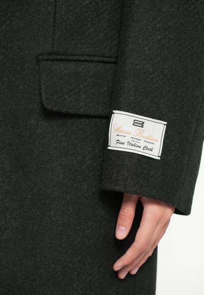 Casaco de lã verde escuro com acabamento texturizado, apresentando um bolso lateral e uma etiqueta com texto em dourado e preto na manga.