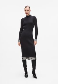 Svart ribbad stickad klänning med hög halsringning, långa ärmar och vita detaljer med "KARL LAGERFELD"-logotyp vid ärmsluten och fållen. Figursydd silhuett.