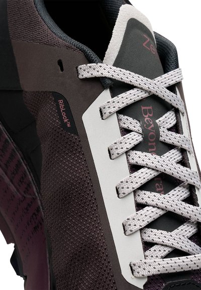 Chaussure de sport marron foncé et grise avec un tissu texturé, des accents violets et des lacets blancs avec un logo sur le côté. Technologie RibLock™ incluse.