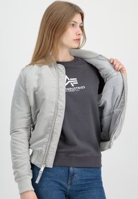 Ljusgrå bomberjacka med en slät, glänsande yta, ribbad krage och ärmslut samt dragkedjestängning. Bärs över en grå sweatshirt.