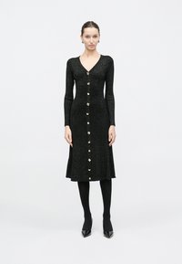 Γυναίκα στέκεται όρθια φορώντας μακρυμάνικο μαύρο πλεκτό φόρεμα με χρυσά κουμπιά μπροστά, μαύρες καλσόν και μαύρα μυτερά παπούτσια.